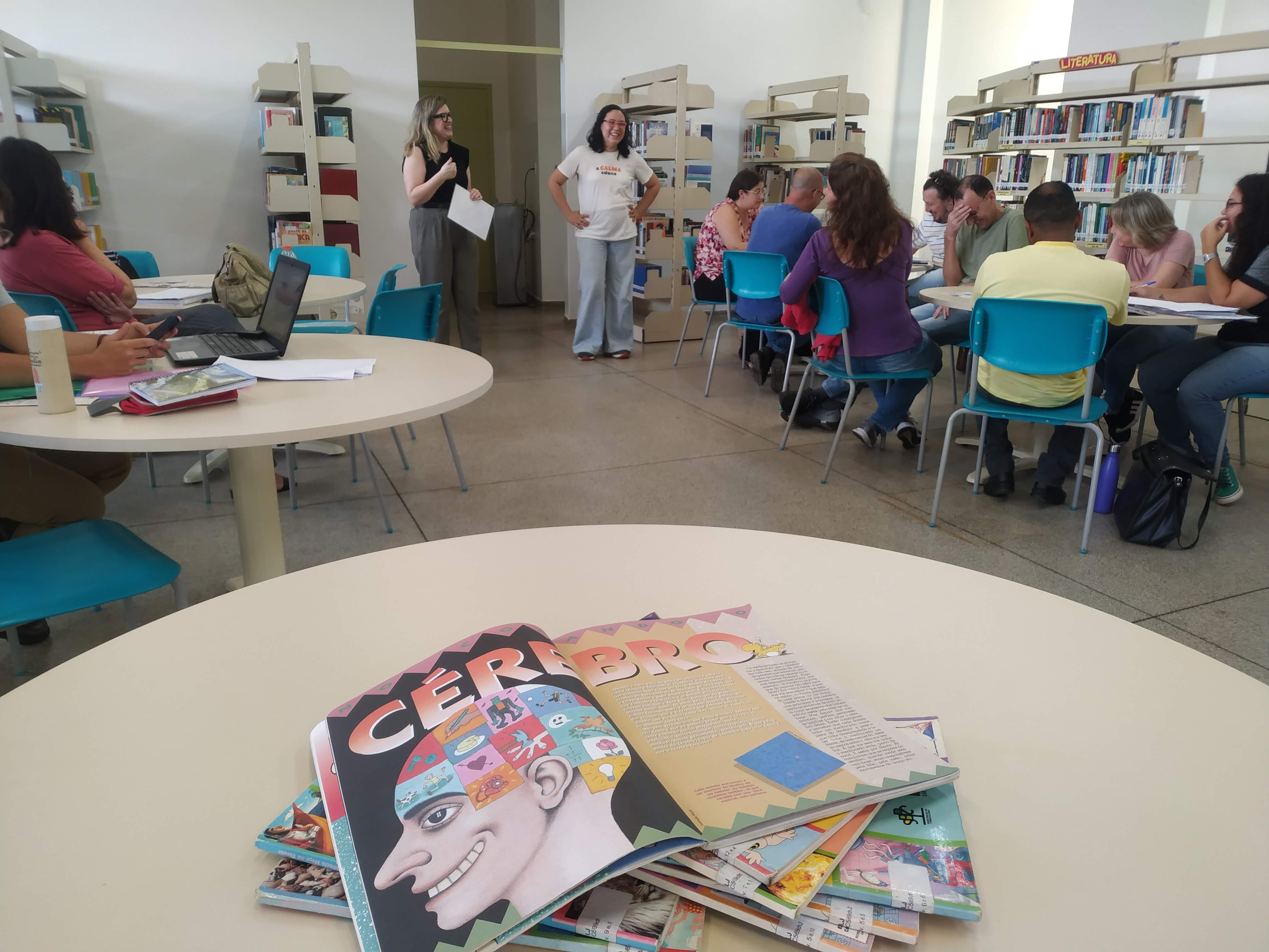 25/03/2024 - Aula do curso piloto do EnvelheCiência, na escola Carmine Botta, na cidade de São Carlos. Aula proferida pela professora Márcia Cominetti e professora Lea Veras, com apoio técnico da mestranda Karen Pulgatti e da comunicadora Giulianna Denari. Na imagem vemos 6 mesas redondas dispostas paralelamente, com professores acomodados entre elas, olhando para as professoras Márcia Cominetti e Lea Veras, enquanto apresentam a proposta do encontro. As professoras Márcia e Lea se encontram ao fundo da imagem, sorrindo, enquanto os professores acompanham o conteúdo. Na mesa mais próxima, vemos um livro didático aberto, com o escrito cérebro em destaque. Foto feita pela comunicadora do LABEN, Giulianna Bueno Denari.