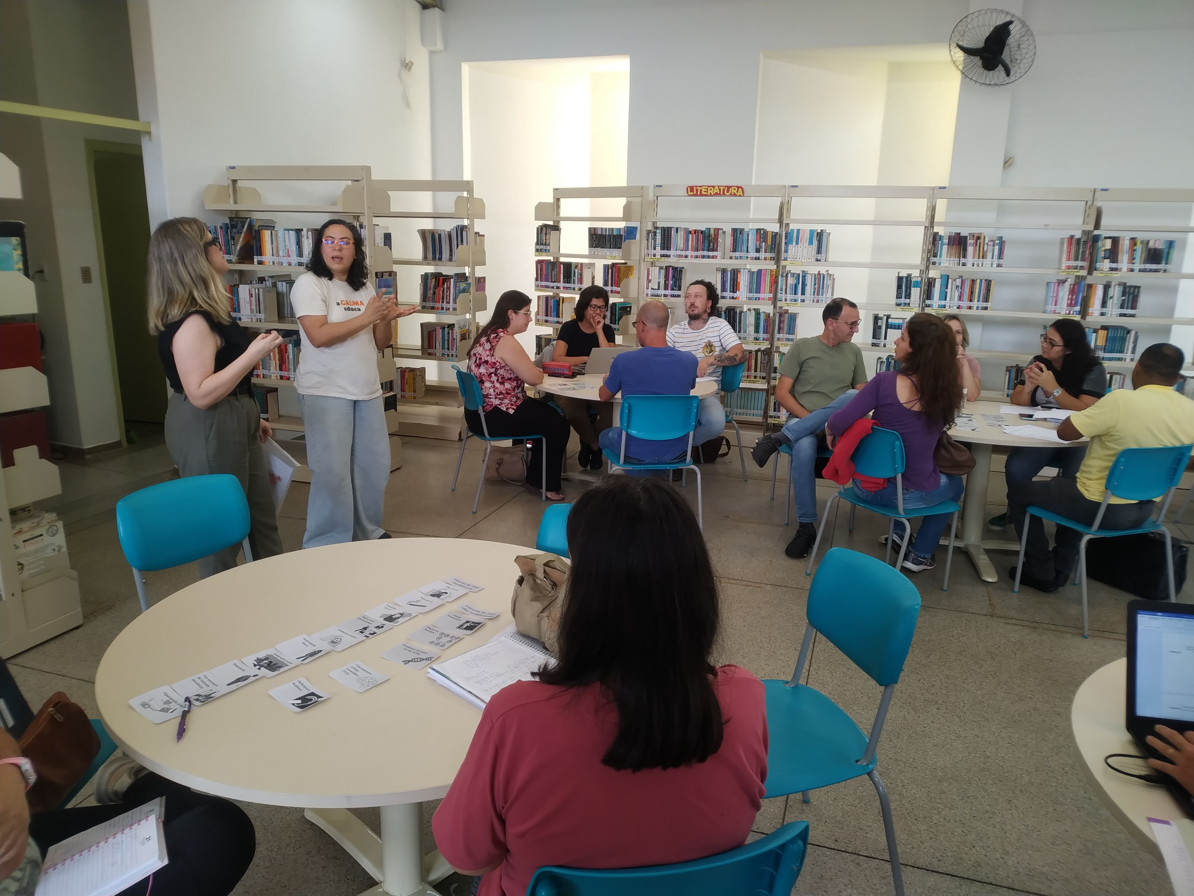 25/03/2024 - Aula do curso piloto do EnvelheCiência, na escola Carmine Botta, na cidade de São Carlos. Aula proferida pela professora Márcia Cominetti e professora Lea Veras, com apoio técnico da gerontóloga Karen Pulgatti e da comunicadora Giulianna Denari. Na imagem vemos 3 mesas redondas dispostas paralelamente, com professores acomodados entre elas, olhando para as professoras Márcia Cominetti e Lea Veras, enquanto apresentam a proposta do encontro. Foto feita pela comunicadora do LABEN, Giulianna Bueno Denari.