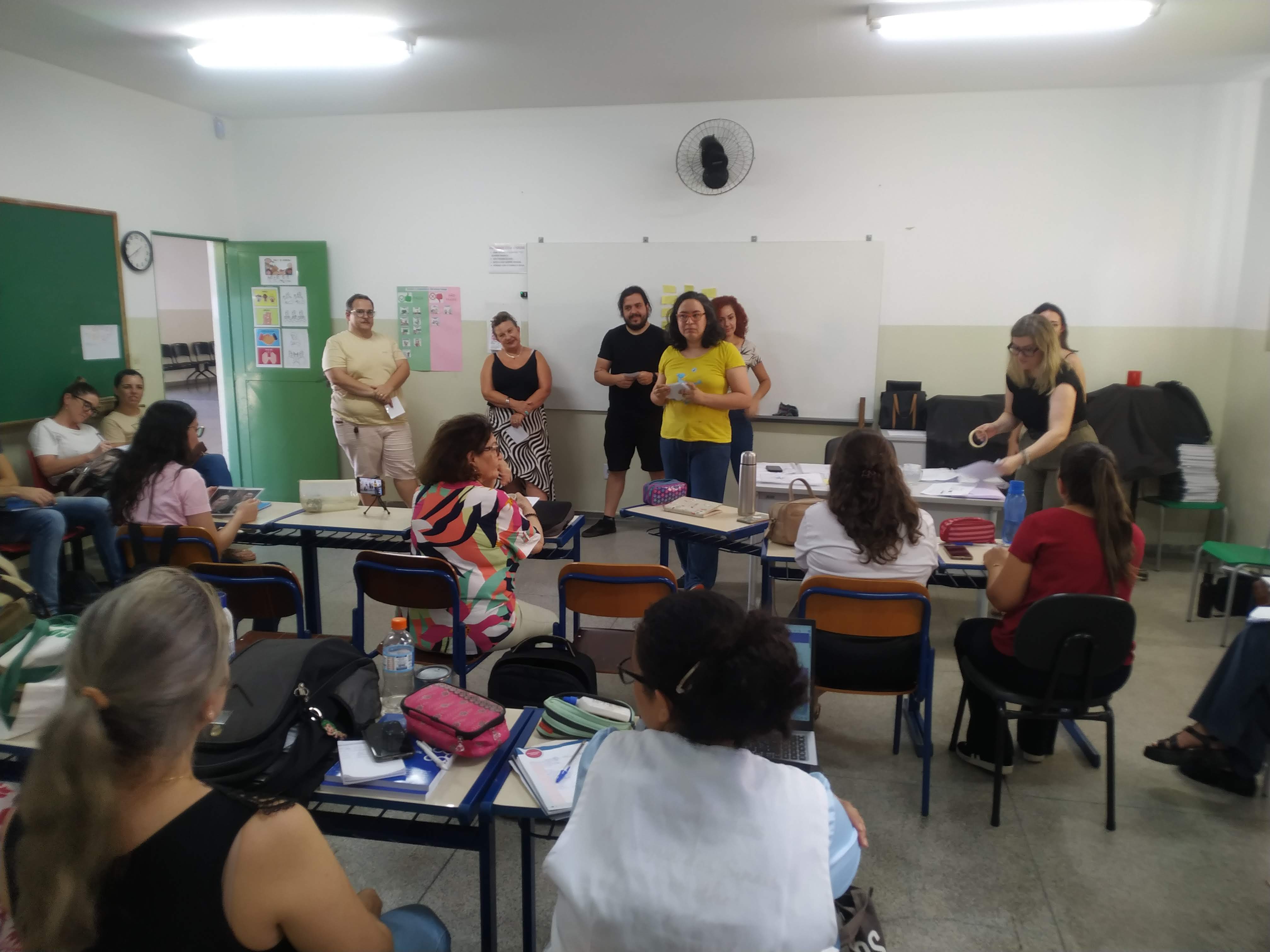 18/03/2024 - Aula do curso piloto do EnvelheCiência, na escola Dalila Galli, na cidade de São Carlos. Aula proferida pela professora Márcia Cominetti e professora Lea Veras, com apoio técnico da gerontóloga Karen Pulgatti e da comunicadora Giulianna Denari. Na imagem vemos os professores sentados em cadeiras e carteiras escolares, de costas, observando a apresentação. À frente da sala, temos professores em pé, participando da dinâmica, junto com as professoras Márcia Cominetti e Lea Veras, bem como a gerontóloga Karen Pulgatti. Foto feita pela comunicadora do LABEN, Giulianna Bueno Denari.