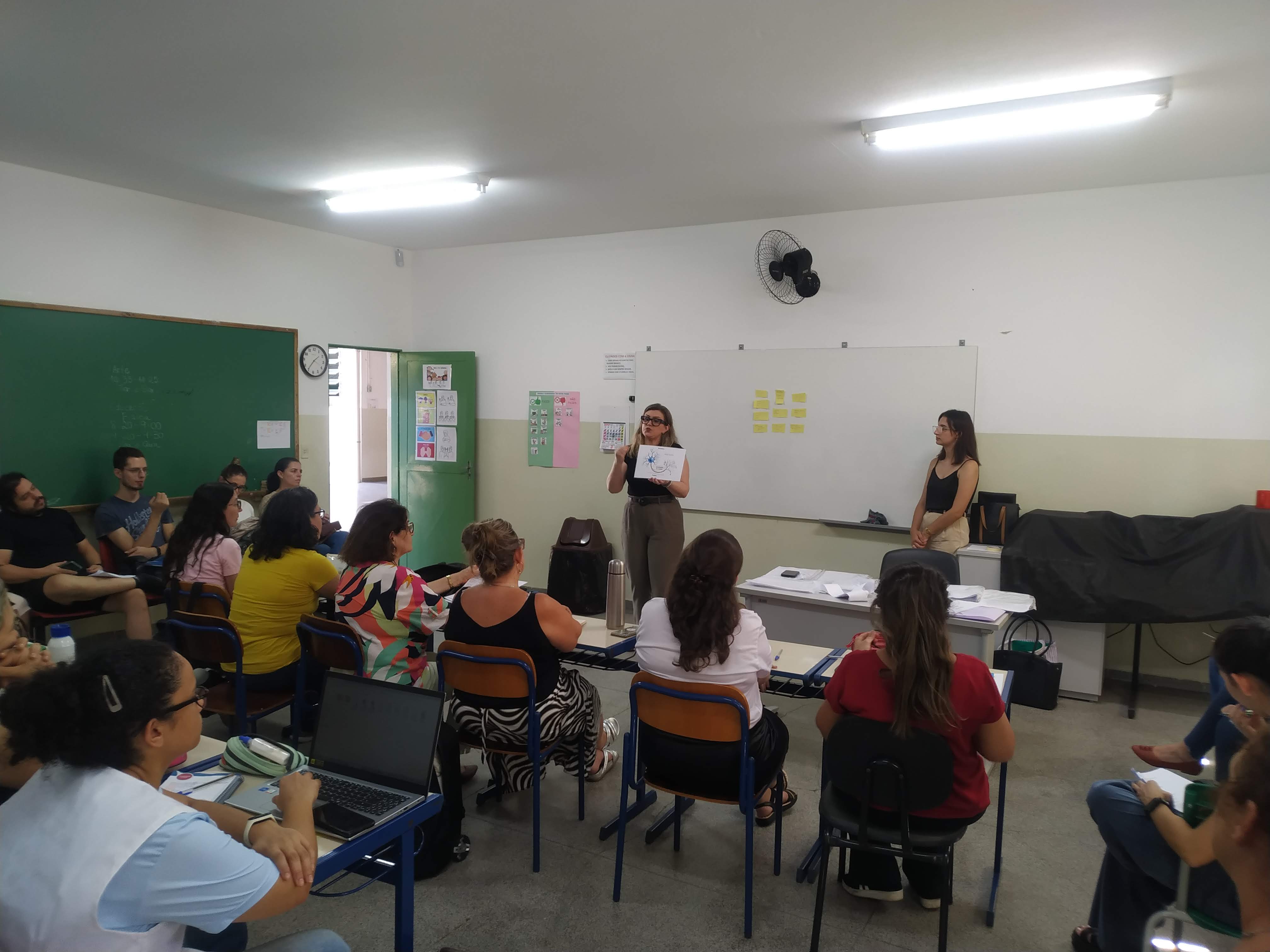 18/03/2024 - Aula do curso piloto do EnvelheCiência, na escola Dalila Galli, na cidade de São Carlos. Aula proferida pela professora Márcia Cominetti e professora Lea Veras, com apoio técnico da gerontóloga Karen Pulgatti e da comunicadora Giulianna Denari. Na imagem vemos os professores sentados em cadeiras e carteiras escolares, de costas, observando a apresentação, enquanto a professora Márcia segura uma imagem ilustrativa e a gerontóloga Karen acompanha a apresentação. Foto feita pela comunicadora do LABEN, Giulianna Bueno Denari.
