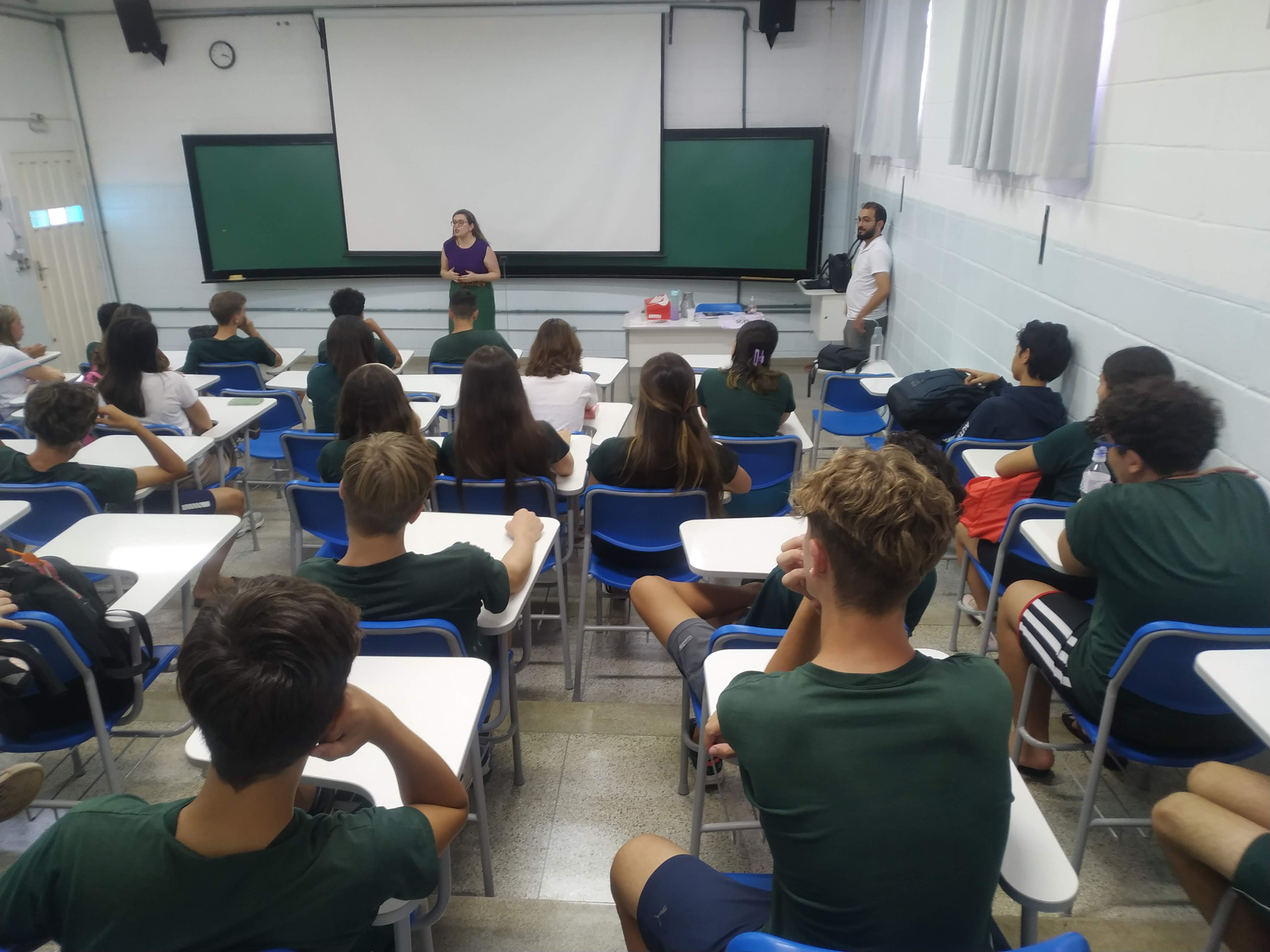 13/03/2024- Palestra proferida pela professora Márcia Cominetti na escola Educativa, em São Carlos, sobre envelhecimento e evidências científicas sobre o funcionamento do cérebro e demências. Na imagem vemos uma sala de aula em desnível, com carteiras de apoio lateral, com os estudantes uniformizados com camisetas verdes, de costas, olhando em direção ao quadro verde ao fundo. A professora Márcia Cominetti, uma mulher branca, de cabelos louros, usando óculos, está vestida de camiseta roxa e calça verde, apresentando a palestra para os estudantes. No canto direito temos o professor João Paulo, coordenador da área de humanas do ensino médio da escola Educativa, assistindo à palestra. Foto feita pela comunicadora do LABEN, Giulianna Bueno Denari.
