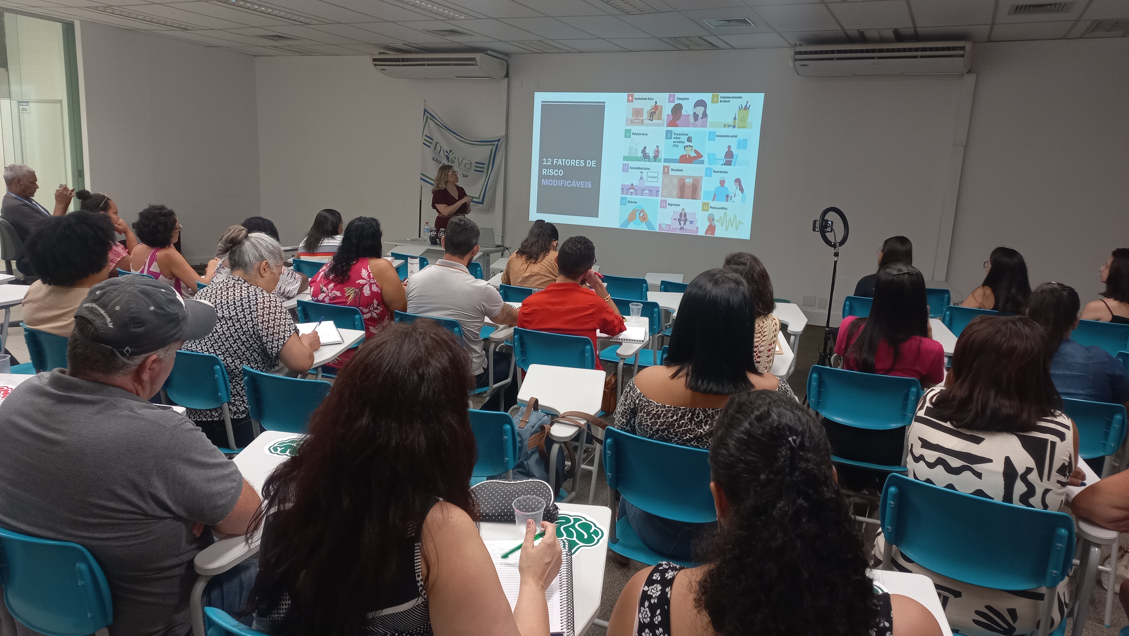 10/01/2025 - Aula do curso piloto do EnvelheCiência, no MOVA, na cidade de São Carlos. Aula proferida pela professora Márcia Cominetti e professora Lea Veras, com apoio técnico das gerontólogas Karen Pulgatti e Letícia Palma. Na imagem vemos a professora Márcia Cominetti, em frente à projeção apresentando os 14 fatores de risco modificáveis para o desenvolvimento de demências, e os professores sentados, de costas, no auditório, assistindo à palestra.